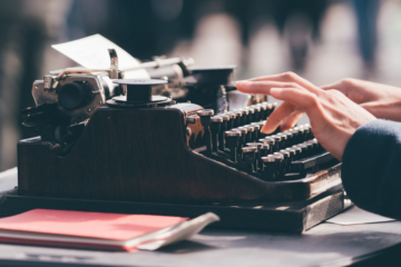 Fingers hover over an old-fashioned black typewriter, with a notebook next to it