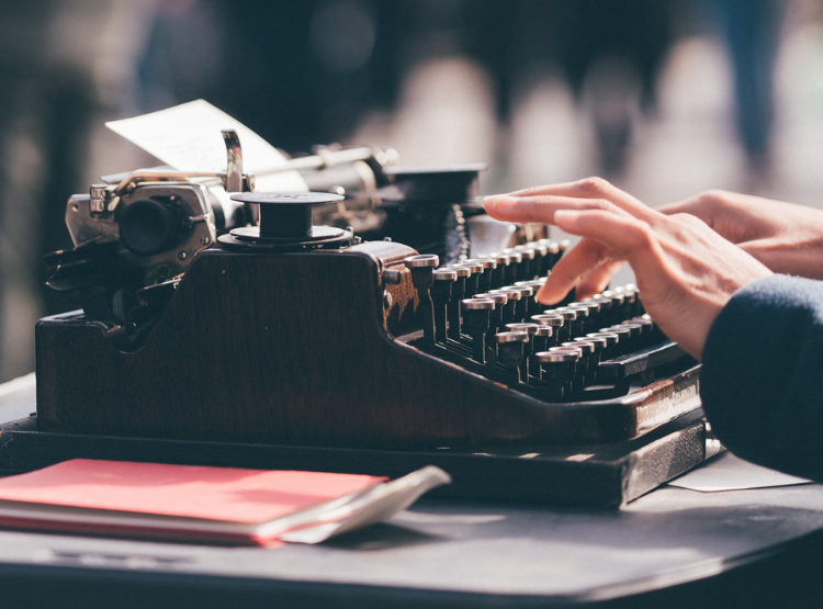 Fingers hover over an old-fashioned black typewriter, with a notebook next to it