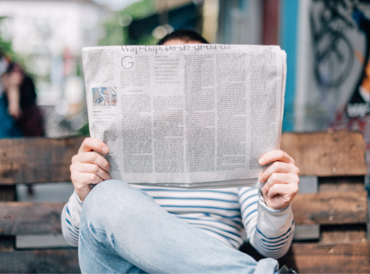 A person sitting on a bench with legs crossed reads a folded newspaper, their face is hidden behind the paper